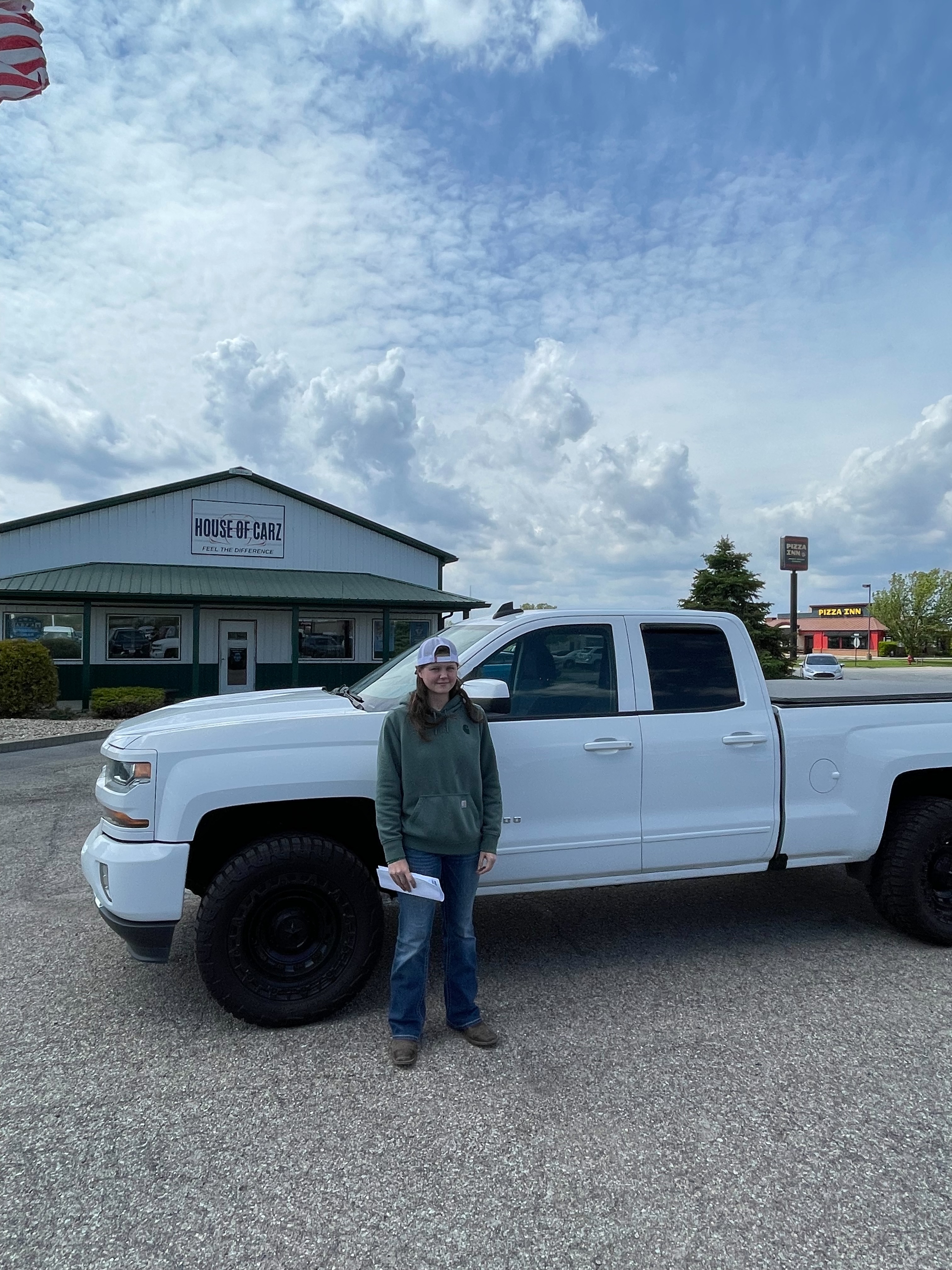 Brianna standing next to her newly-purchased 2018 Chevrolet Silverado in front of the House of Carz dealership in Rochester, Indiana, holding the paperwork from her purchase on May 9, 2026.